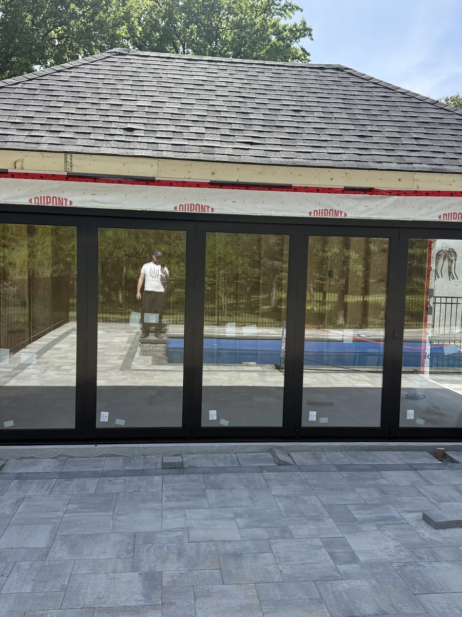 The pool cabana's wall of black-framed glass panels during installation, pool reflected in the glass